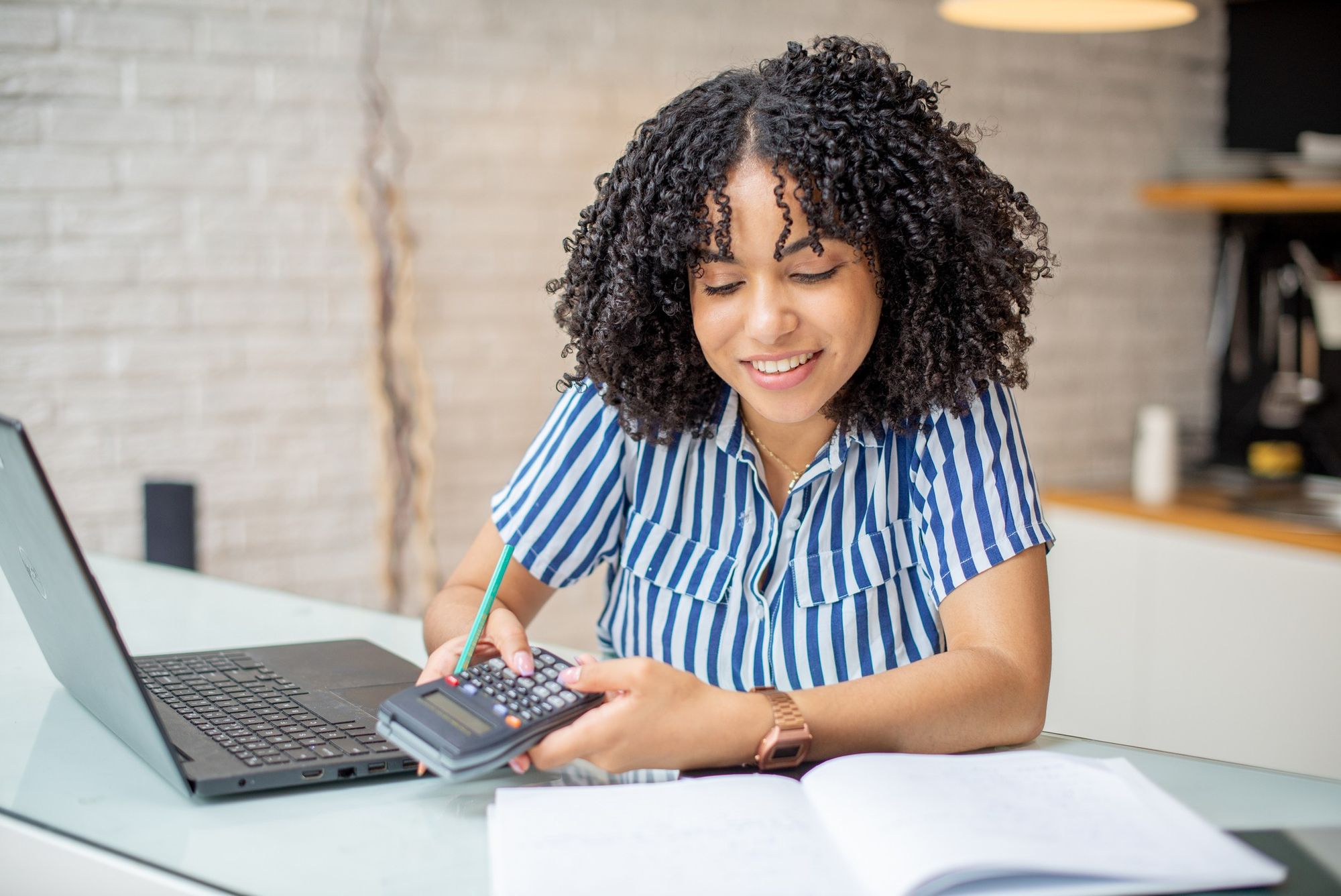 Mulher com uma calculador na mão olhando para um caderno e com um notebook ao lado dela